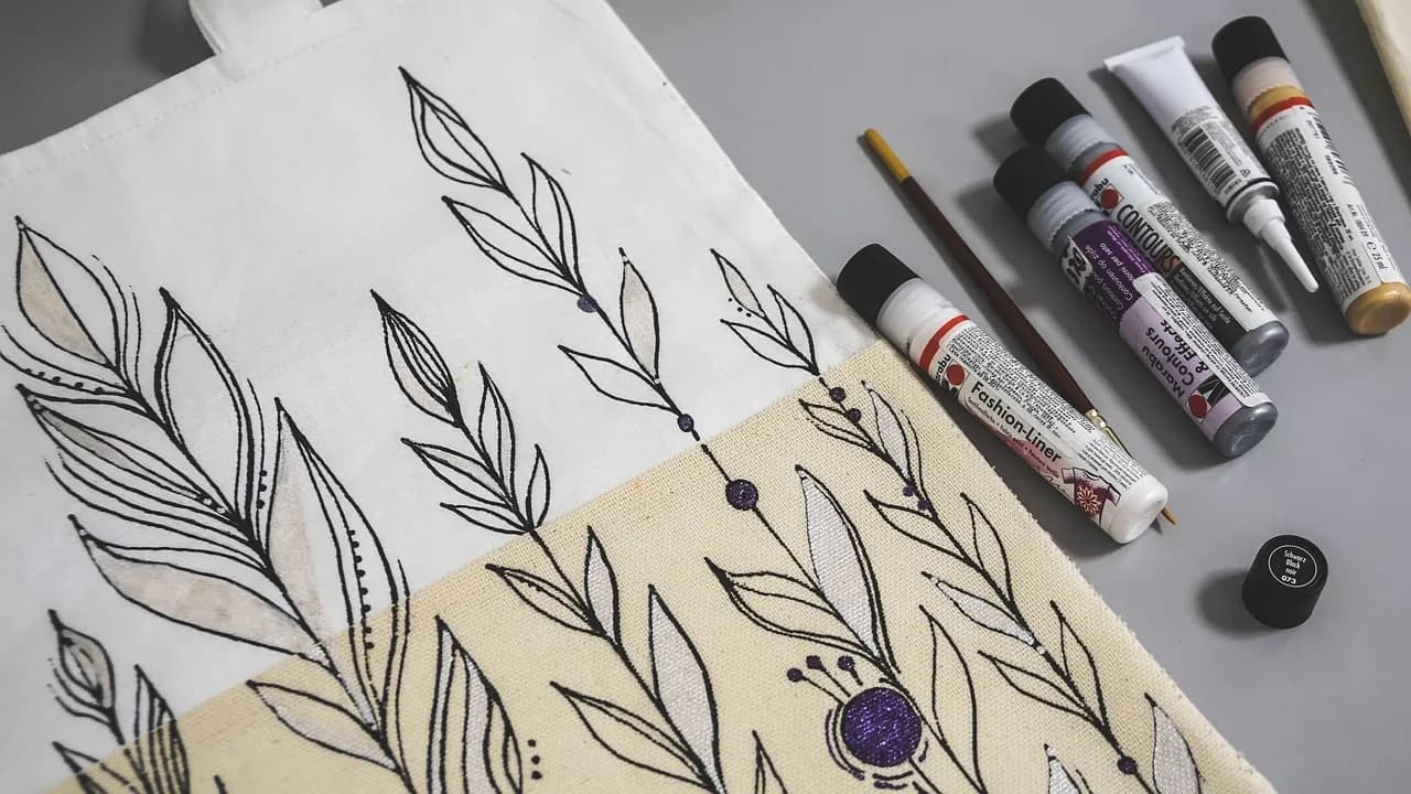 Hand-decorated canvas tote bag with a leaf design, laid out beside fabric paint tubes